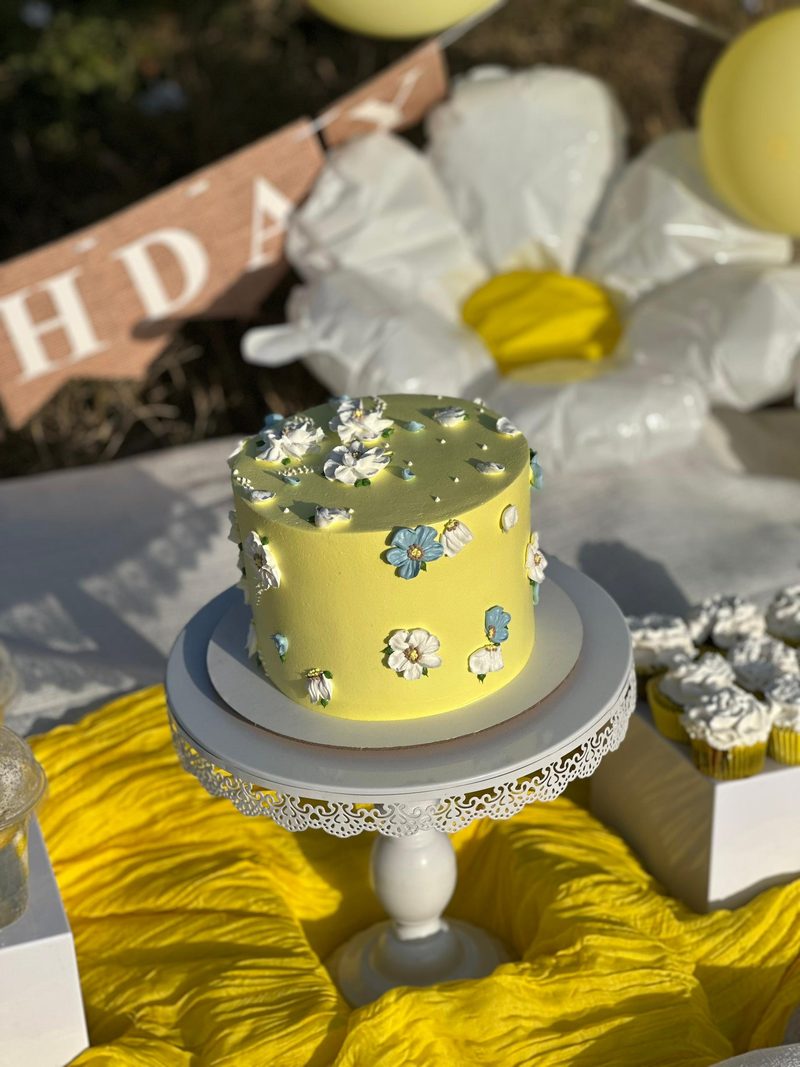 كيكة أقحوان من Bella Chef في تعز: كيكة صفراء صغيرة مزينة بزهور أقحوان بيضاء وزرقاء على حامل أبيض